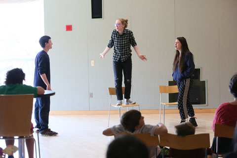 Three students, one of which is standing on a chair, acting a scene for festival attendees.