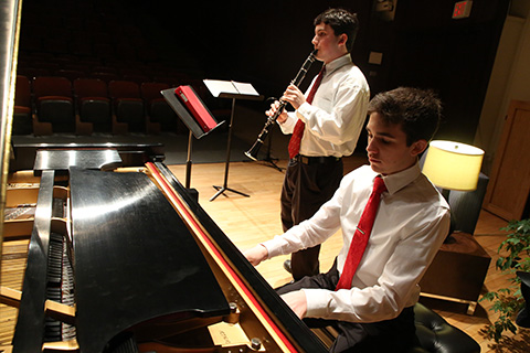 One person playing the piano in front of another person playing the clarinet.