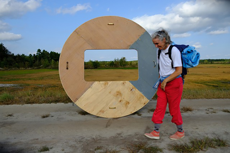 Tory Fair outdoors, windswept, with a large plywood wheel with a rectangle cut out of the center