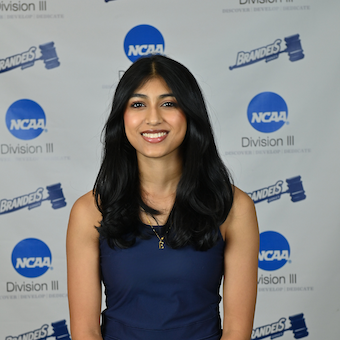 Ella smiles, wearing a blue tank top and gold necklace, standing in front of a Brandeis sports backdrop.