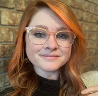 Jessi Brewer, pictured with auburn hair and glasses, smiles at the camera.