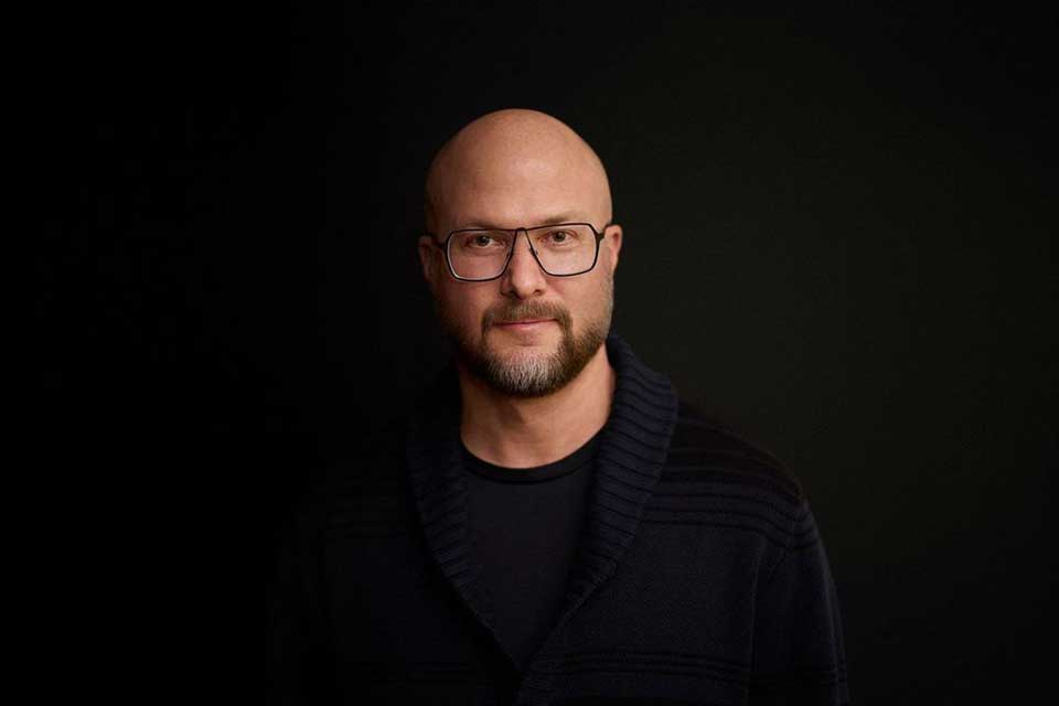Portrait of  Trent Preszner wearing rectangular glasses and a dark sweater over a black shirt. He faces the camera with a neutral expression against a solid black background with soft studio lighting.