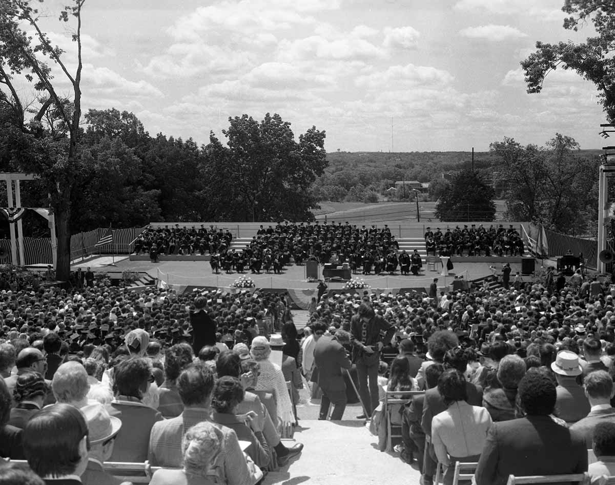 Black and white photo of a Commencement ceremony outdoors on the Brandeis campus. Graduates sit on stage while spectators are seated in the audience.