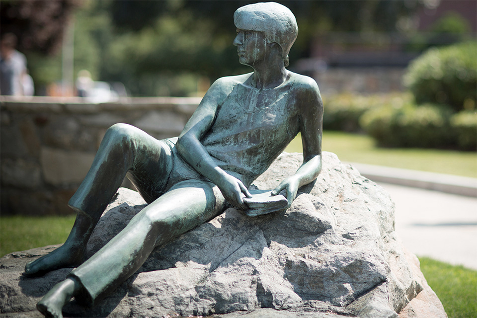 A bronze figure seated on a rock holding a book gazing out into the distance