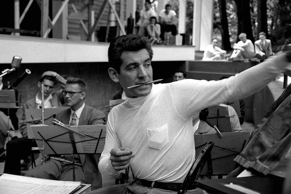Black and white photo of Leonard Bernstein conducting at Brandeis. He holds a pencil in his mouth.