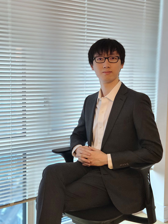 Licheng Shen has on a suit and glasses. He is sitting in front of a window with blinds. His legs are crossed and he is gazing directly at the camera.