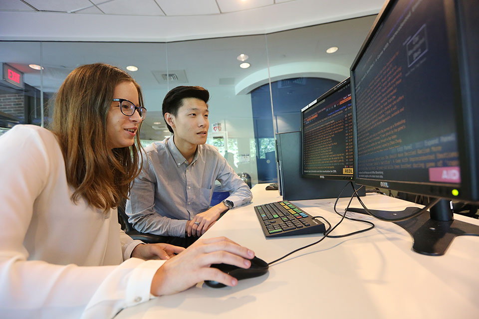 Two students, one using a computer mouse, look at two computer monitors. 