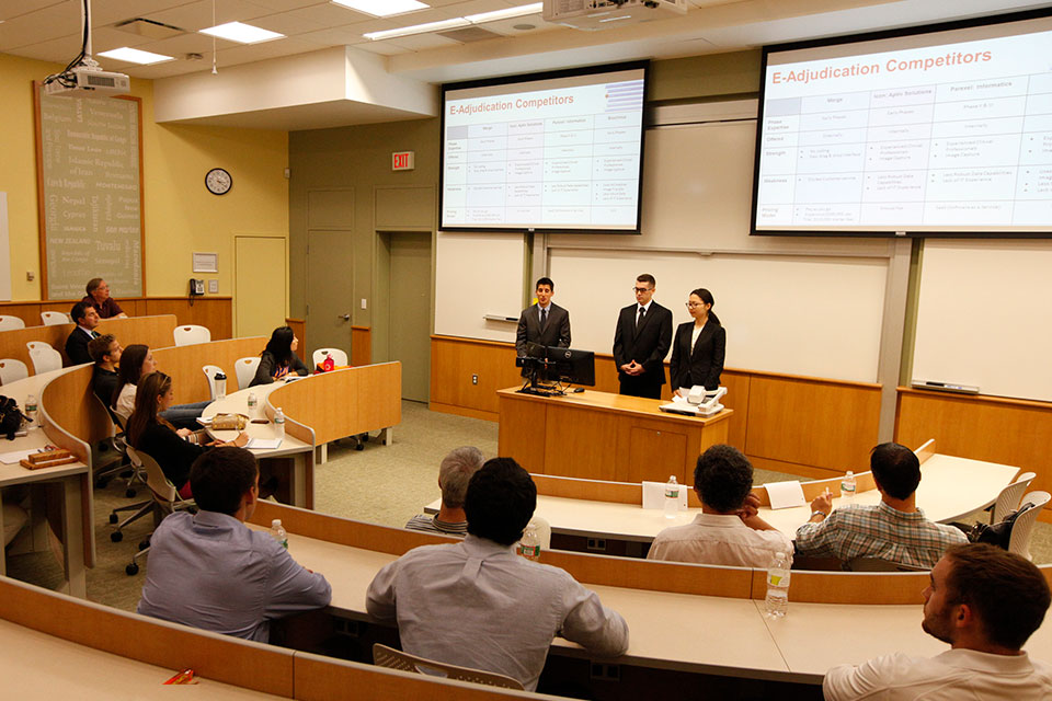 Three students in suits do a presentation at the front of the room.