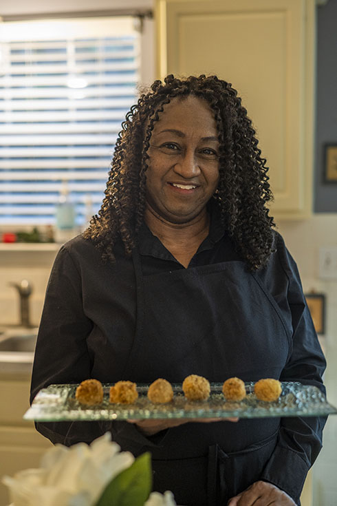 Dawn stands holding a tray of appetizers, smiling and wearing a black chef's jacket.
