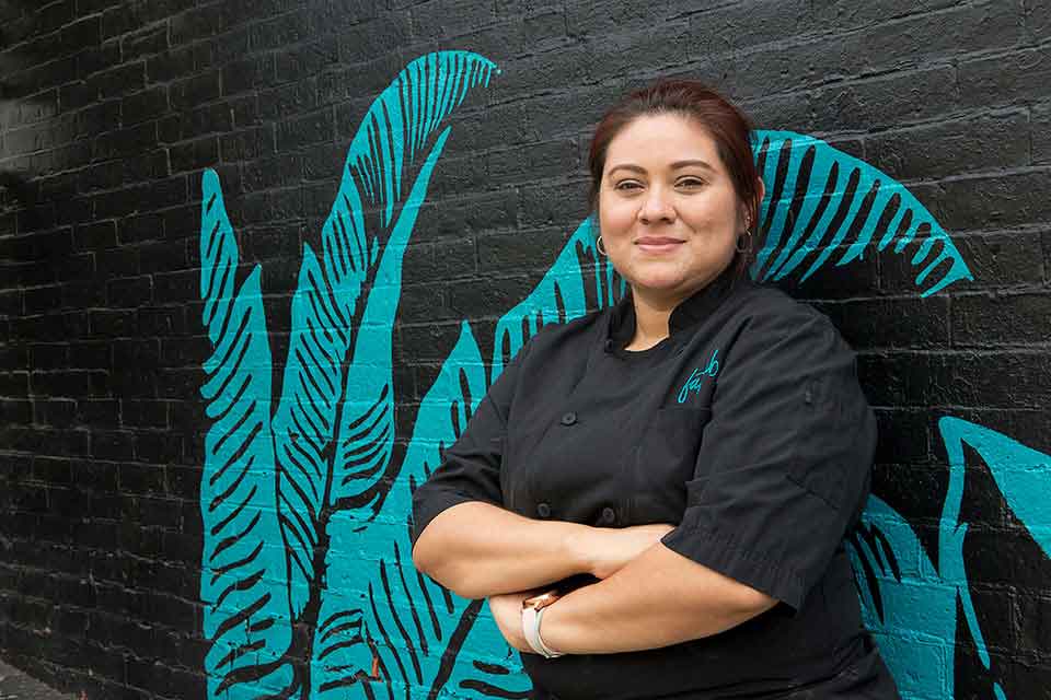 Mayra, wearing a chef's coat, stands against a black wall with a teal plant painted on it.