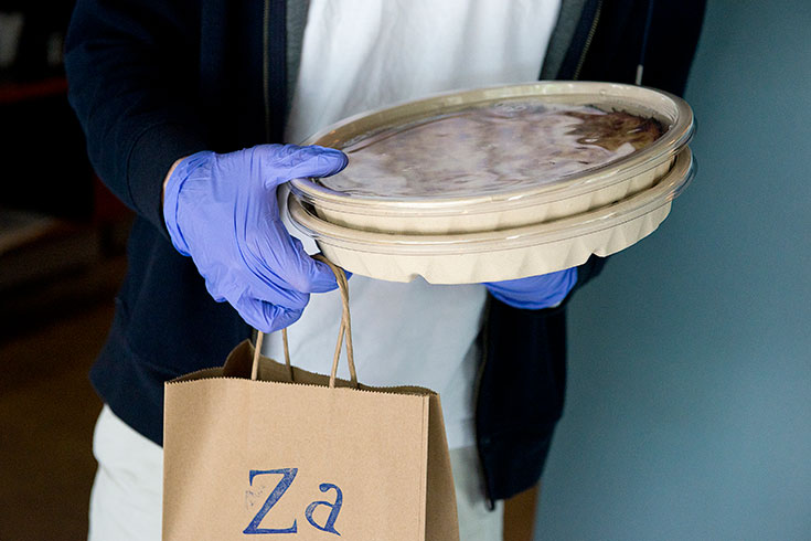 A person wearing gloves stands holding 2 pizzas in takeout containers