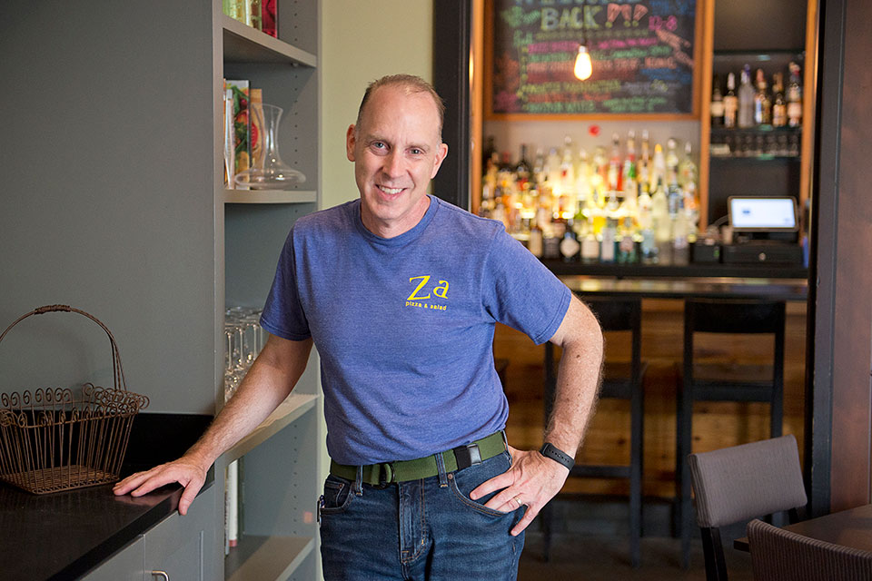 Steve stands in a restaurant wearing a Za restaurant shirt