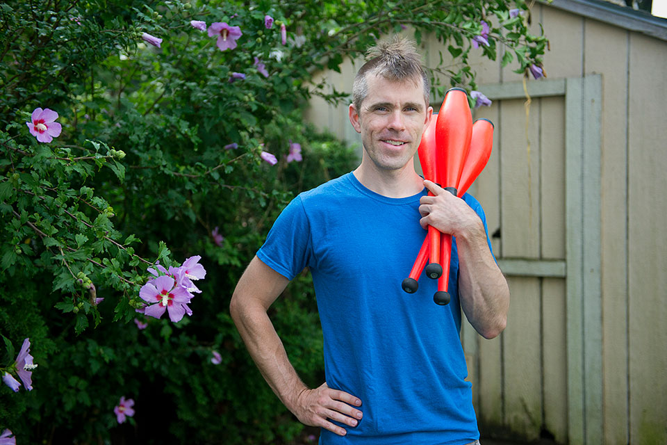 Tim stands in front of a flowering bush holding three juggling clubs
