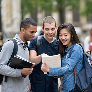 3 students standing with backpacks on looking at a book