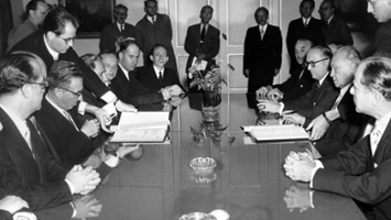 Group of men in suits sitting at a big table with paperwork in black in white