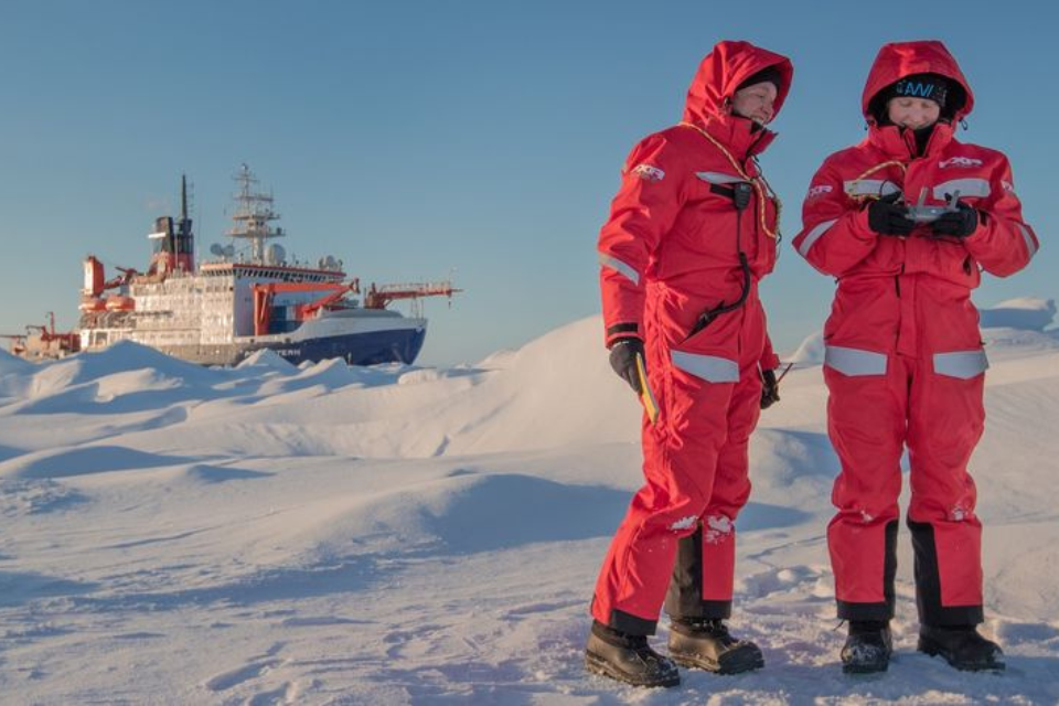 Two people on ice in orange suits with a ship in the background