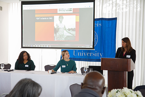 Jennifer Miller speaking at podium accompanied by slides during a panel discussion. Seated panelists to her left are Cynthia Porter and Diana Filar.