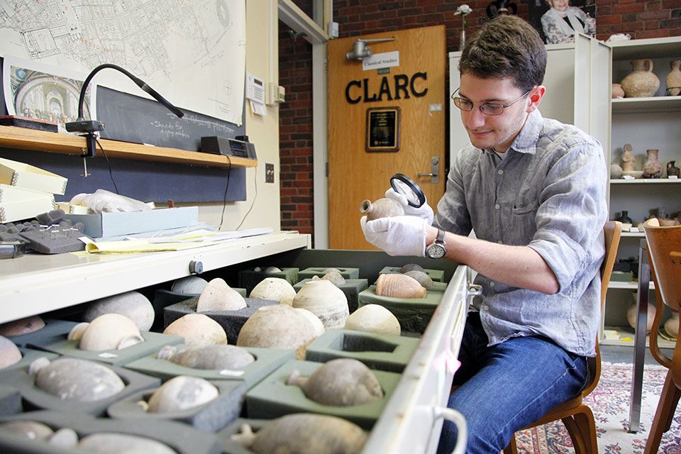 A student looks at a stone vase with a magnifying glass, while sitting next to a drawer filled with more stone vases.
