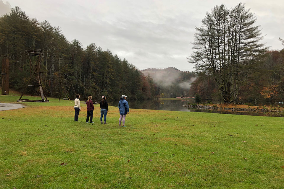 Four people standing on grass looking toward water and the woods