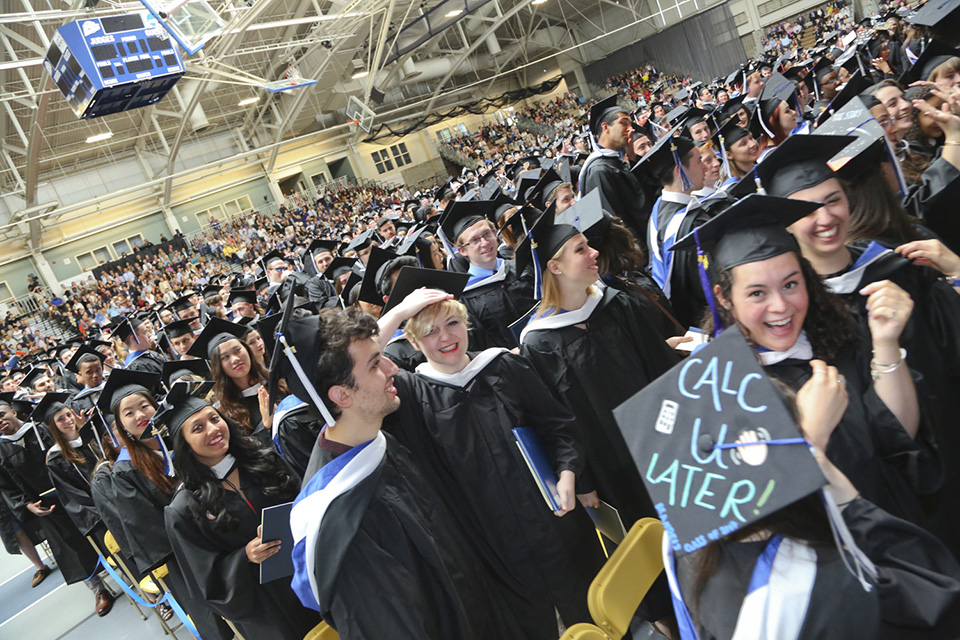 A happy crowd of accomplished Brandeis undergrads anxiously awaits what the future has in store.  A student in the front has a mortarboard with these words written on it: Call U Later and a picture of a cell phone