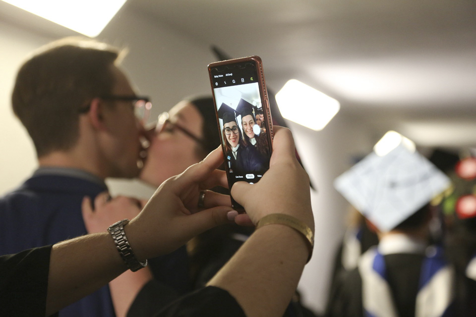 Two students kiss in the background while another takes a photo with a cell phone