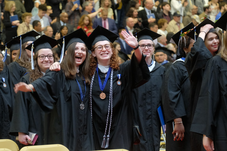 Students in caps and gowns wave to others in the crowd