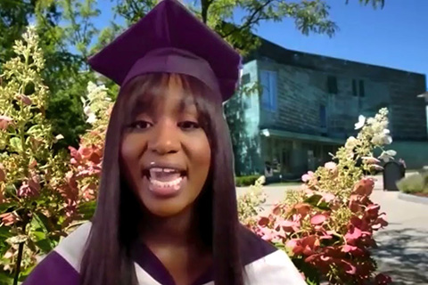 Jainaba Gaye wearing a graduation cap and gown with the campus center and flowers in the background 