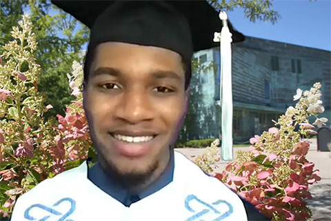Kwesi Jones wearing a graduation cap and gown with the campus center and flowers in the background
