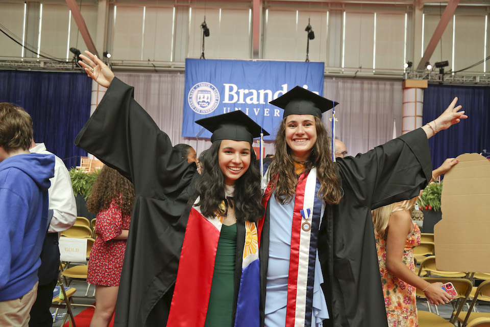 Two graduates stand side-by-side, smiling with their hands in the air.
