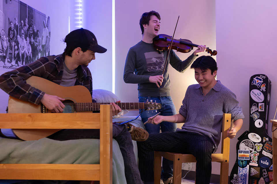 Students play a variety of string instruments in a dorm room and laugh.