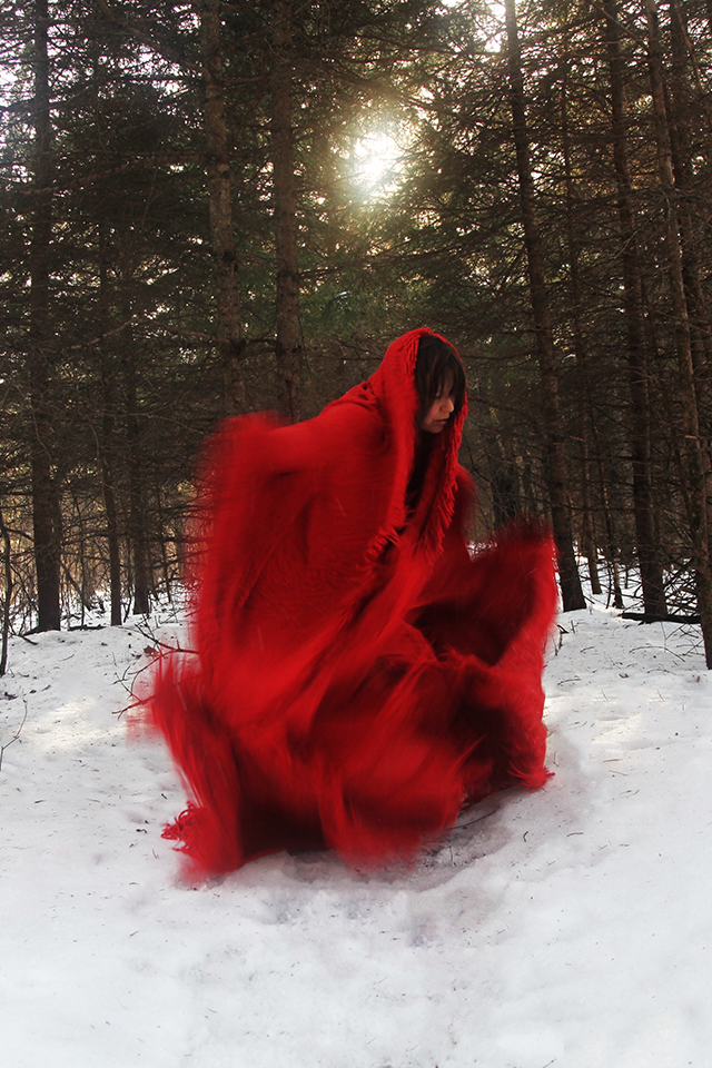 A person wrapped in a large, flowing red fabric stands in a snowy forest, partially bent forward, with the cloth billowing around them in motion. Sunlight filters through the tall trees in the background, creating a dramatic and ethereal scene.