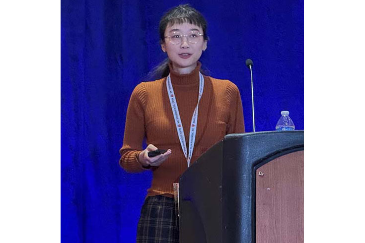 Jin Zhao giving a presentation next to a wooden podium. The background is blue. She is holding a remote in her right hand