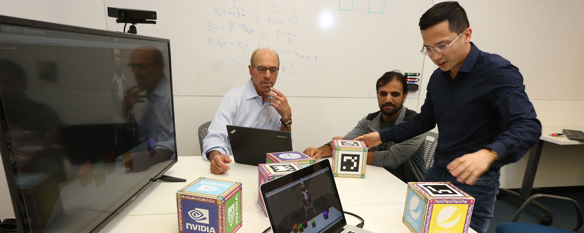 A professor sits with two students in a computer lab setting.