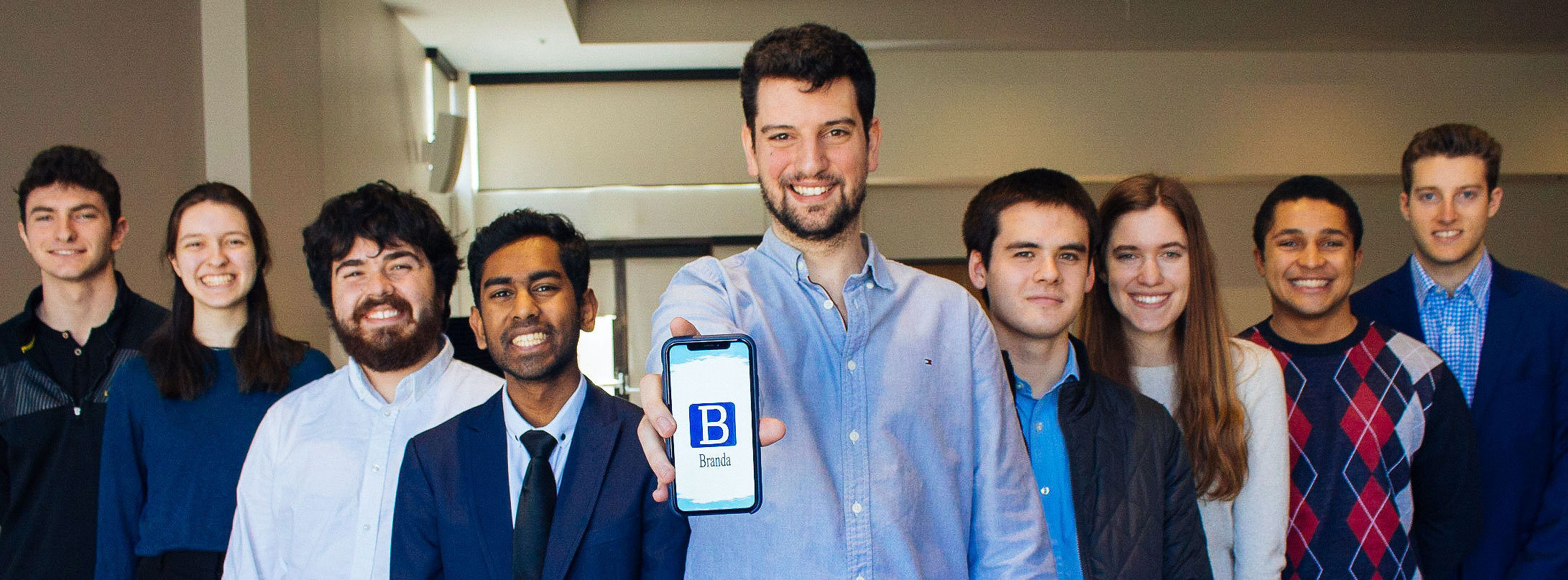 A group of students all smiling at the camera while the student in front holds up a phone with the Branda app displaying on it
