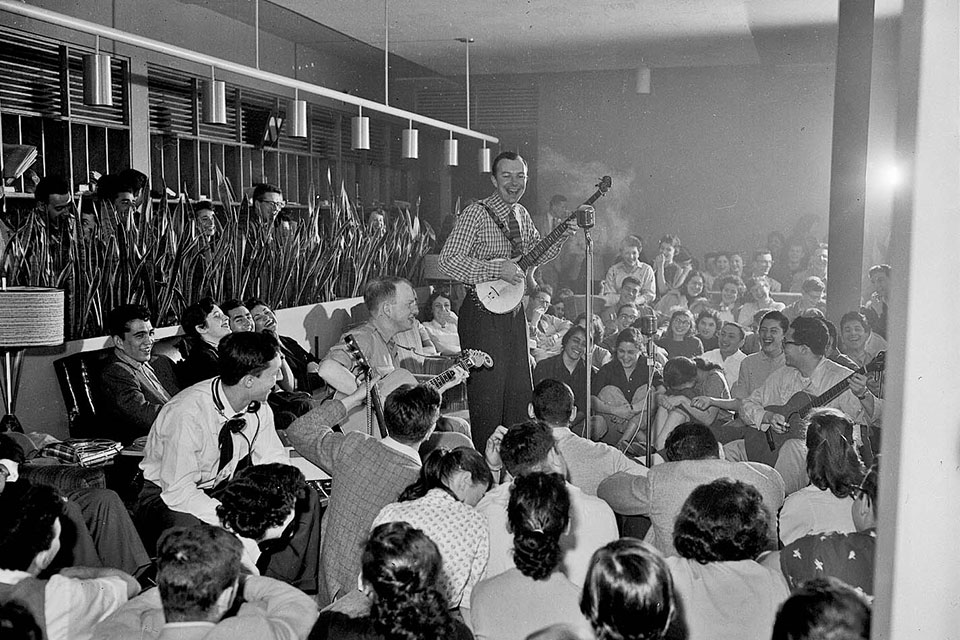 Pete Seeger plays banjo on a stage surrounded by students sitting on the floor