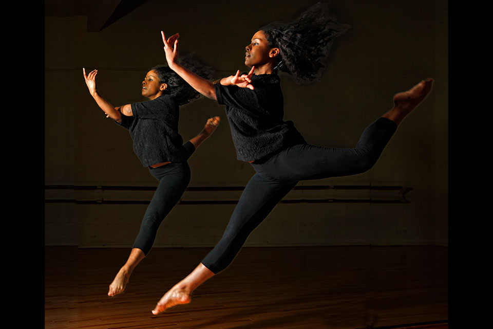 A dancer wearing all black leaps in front of a mirror