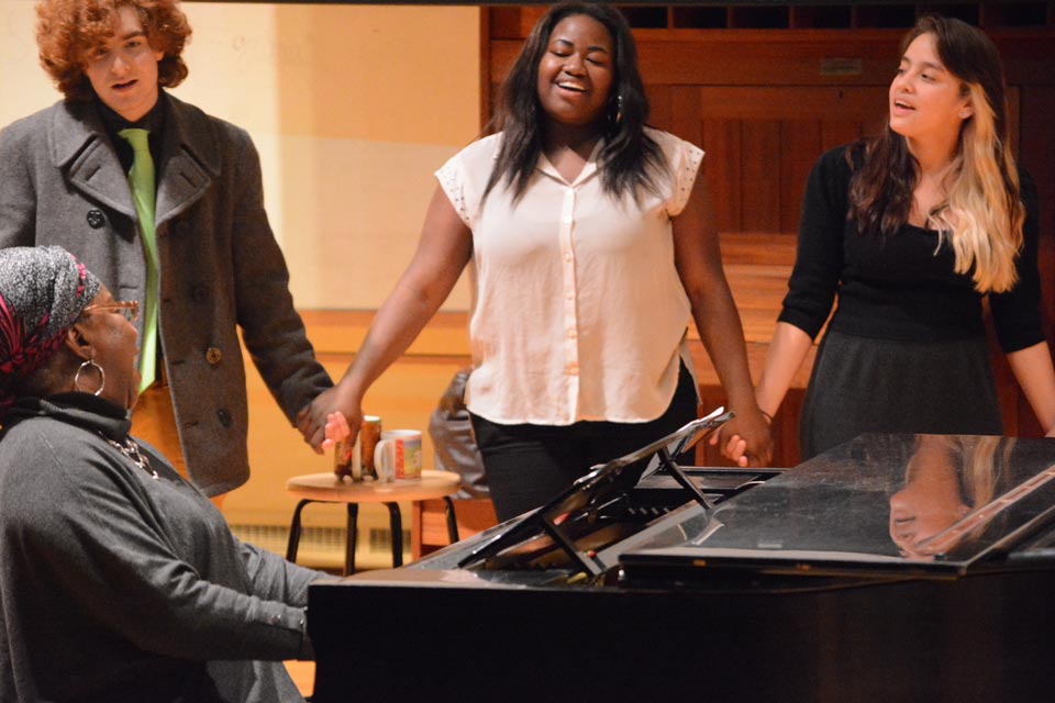 students holding hands and singing around a woman playing the piano
