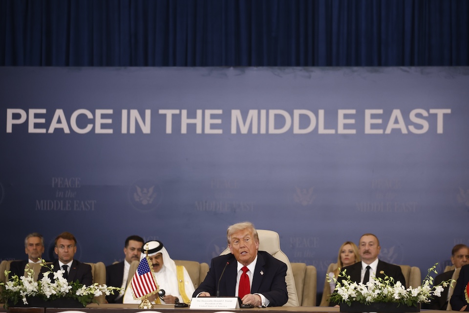 President Donald Trump sits at a table at the ceasefire signature ceremony, behind him international leaders, behind them a banner reading "Peace in the Middle East."