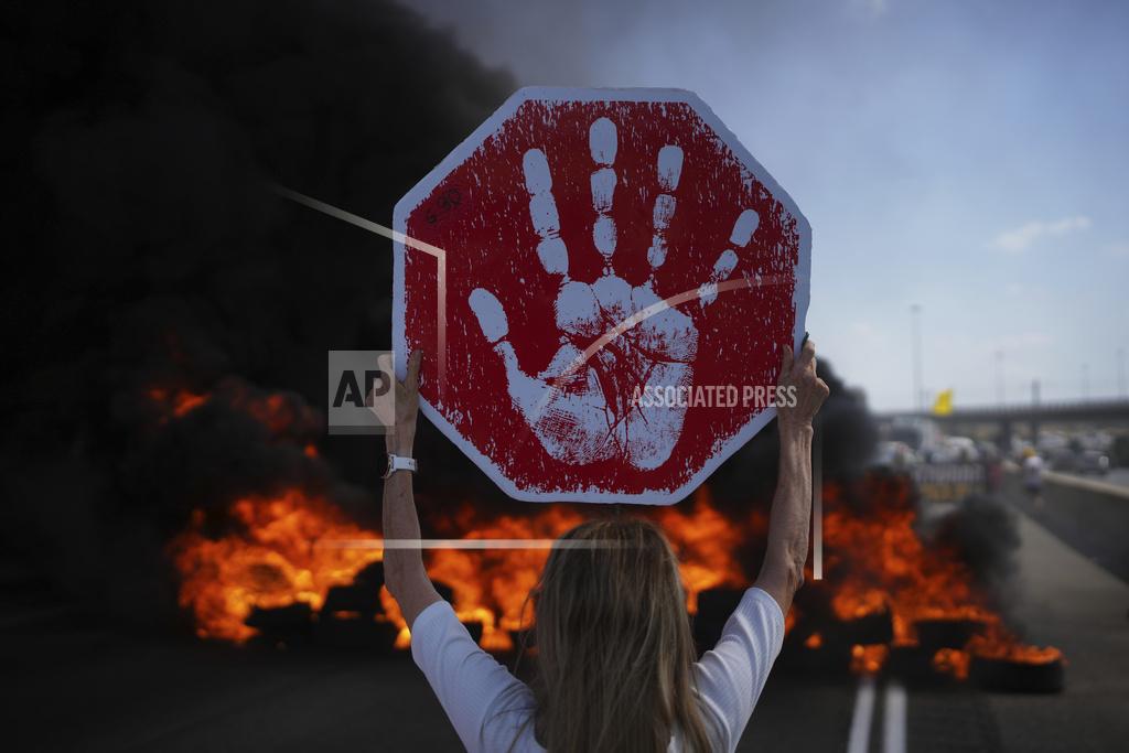 A protestor holds up a sign with a handprint on it, blocking a highway with smoke in the background.