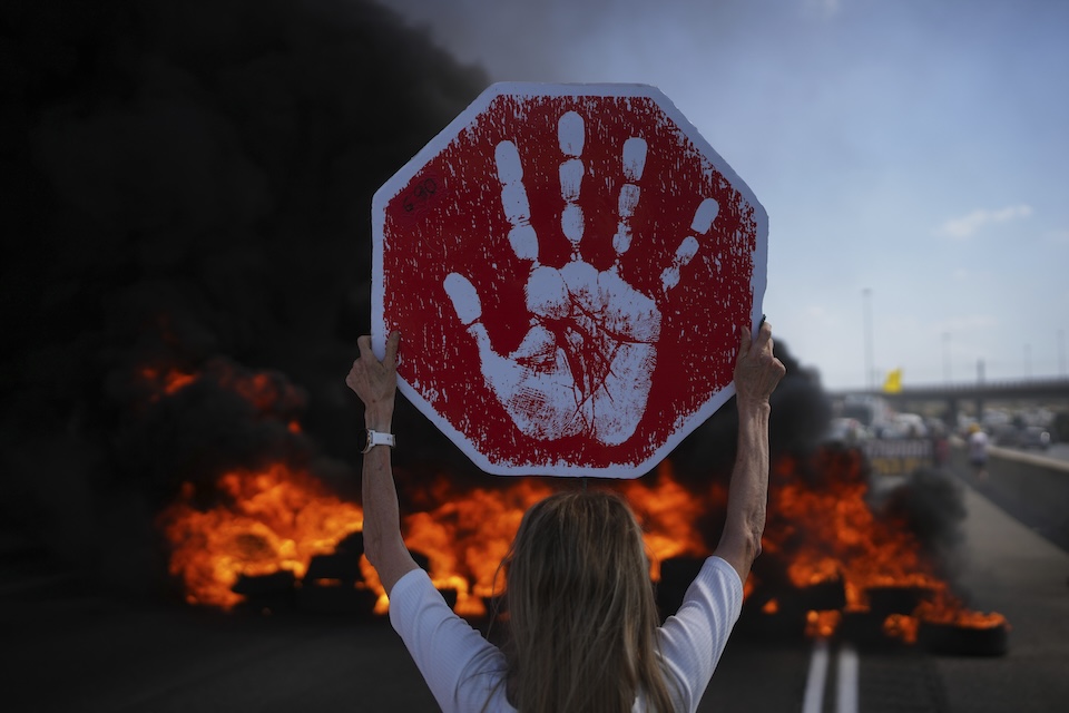 A protestor holds up a sign with a handprint on it, blocking a highway with smoke in the background.