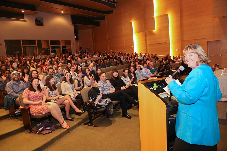 Stephanie Grimes at podium in front of filled auditorium