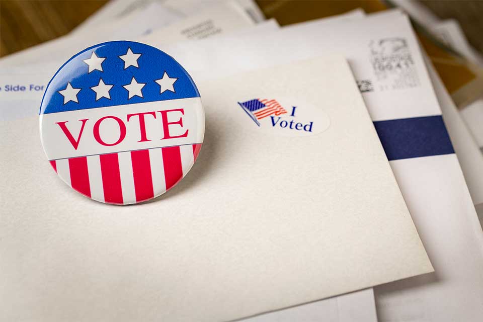 Picture of a vote pin and an I voted sticker on top of 2 envelopes