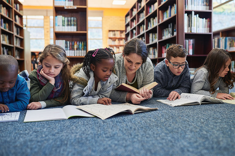 A teacher, center, among elementary school students, reading books in a library on the floor