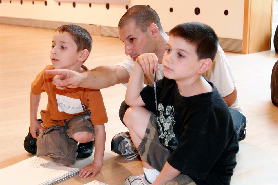 A teacher sitting with two young boys, pointing to something beyond the image.