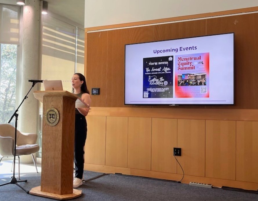 Nicole Lockhart-Rios speaking behind a podium next to a projector screen showing Upcoming Events