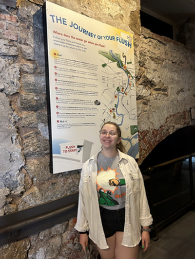 Andie stands by a stone archway in front of a sign that reads “The Journey of Your Flush” at the Fairmount Water Works in Philadelphia