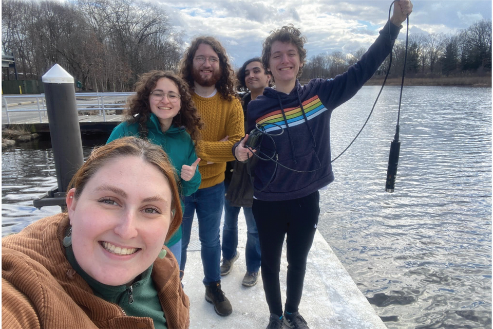 Students standing at the Charles River with a meter to measure