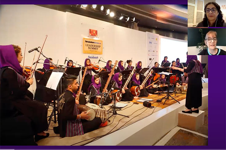 A video still of an orchestra playing with two people on Zoom in the corner