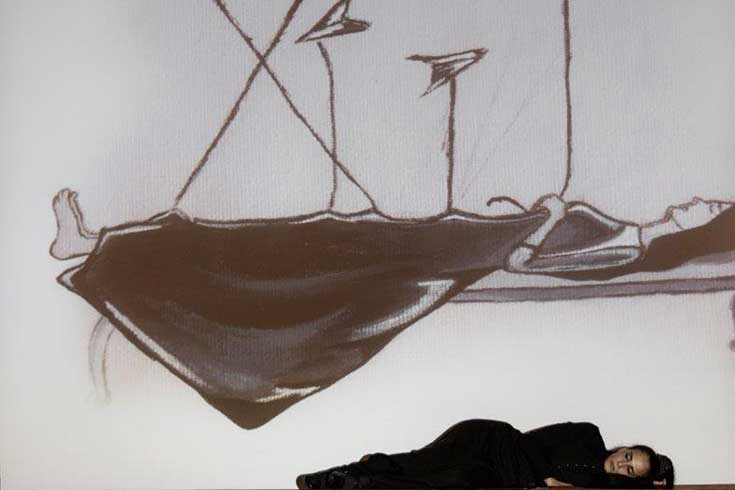 A black and white drawing of a woman laying on platform with airplanes flying around while an actual woman lays on the floor below.