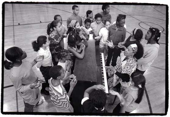 Children surround Jane Sapp while she is playing piano